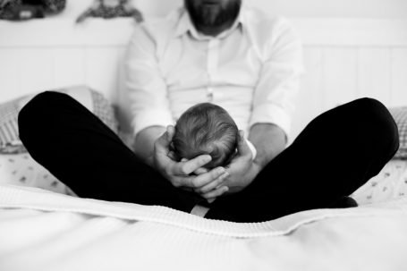 Vater hält ein neugeborenes Baby in den Händen, auf einem Bett sitzend.