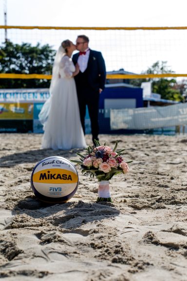 Brautpaar küsst sich am Strand, ein Volleyball und Blumenarrangement im Vordergrund.