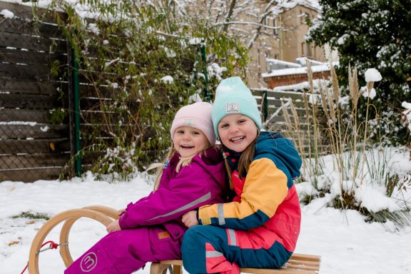 Zwei Kinder im Schnee sitzen auf einem Schlitten und lächeln.