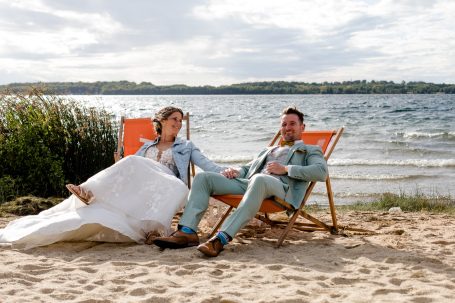 Brautpaar sitzt entspannt auf Liegestühlen am Strand, im Hintergrund einsee und Bäume.