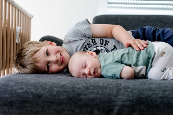 Ein lächelnder Junge sitzt mit einem schlafenden Baby auf einer Couch.