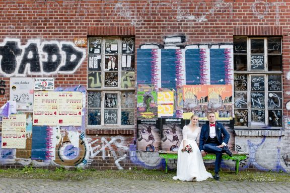 Hochzeitspaar sitzt auf einer Bank vor einer graffiti-bedeckten Backsteinwand.
